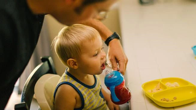 Niño pequeño sentado en la mesa sosteniendo una taza con sorbete y papá parado detrás de él