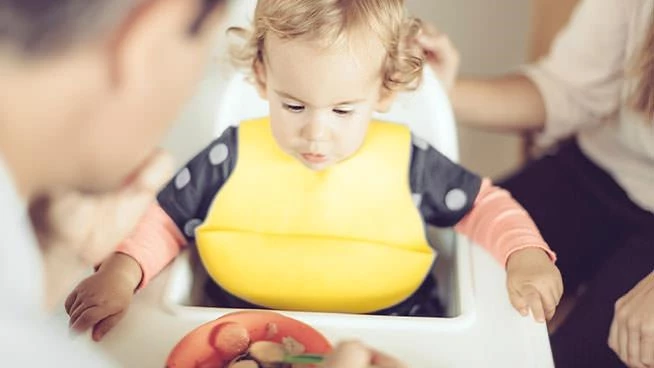 Niño sentado en una silla alta comiendo comida con sus padres sentados cerca