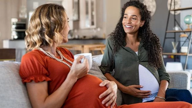 Two pregnant women sitting on the couch talking with one holding a cup of coffee