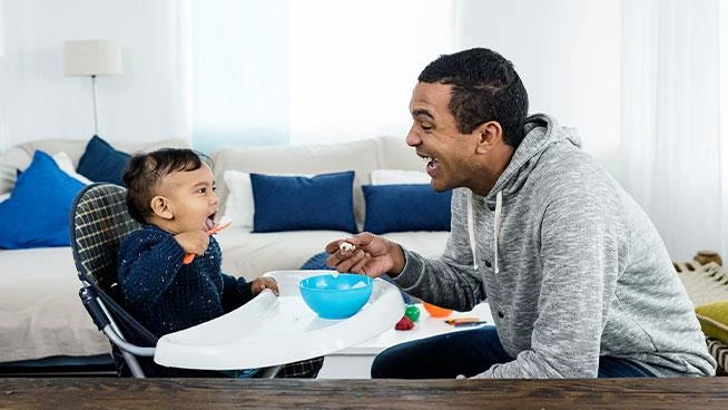 Toddler son in a high chair being fed yogurt by dad