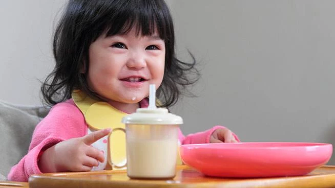 Niña pequeña feliz sentada en la mesa con su plato y taza para sorber