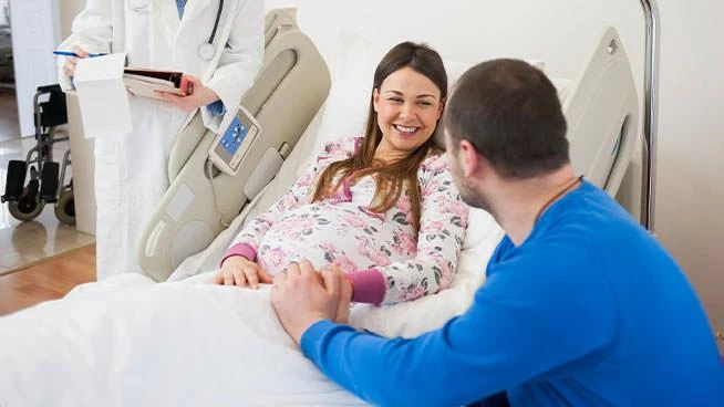Mujer embarazada acostada en una cama de hospital sonriendo mientras su cónyuge le toma la mano al médico al fondo