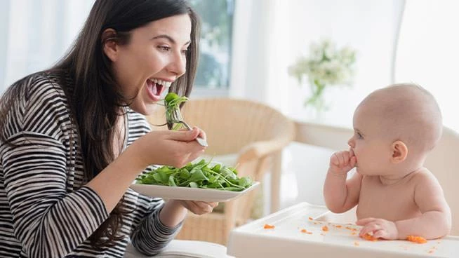 Mom eating salad while baby eats pieces of carrot in high chair