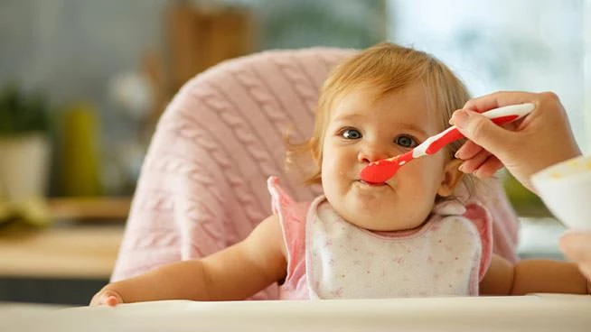Parent spoon feeding infant daughter as she sits in a high chair