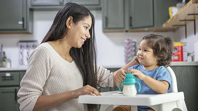 Mom smiling at baby in a highchair who has a milk bottle